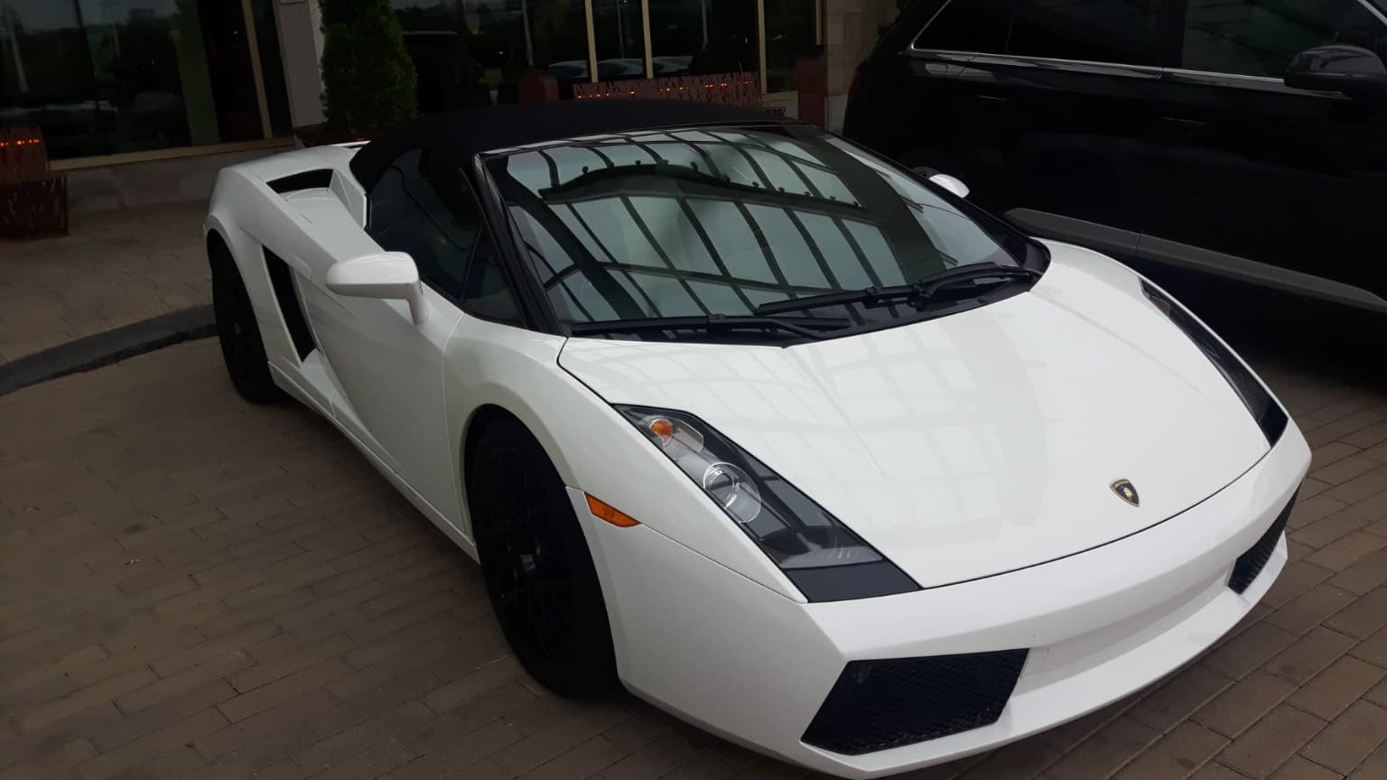 White Lamborghini convertible with black wheels and a black soft top parked on brick pavement.